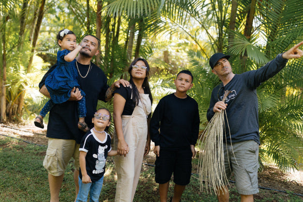 A family of five stands together in a lush tropical garden with palm trees. The father on the left holds a baby girl, while another young boy stands in front of them.