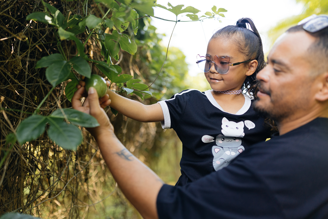 A young girl wearing a black graphic t-shirt and sunglasses stands outdoors next to a man, reaching toward a green fruit on a leafy vine. The girl's shirt features a white cat graphic.